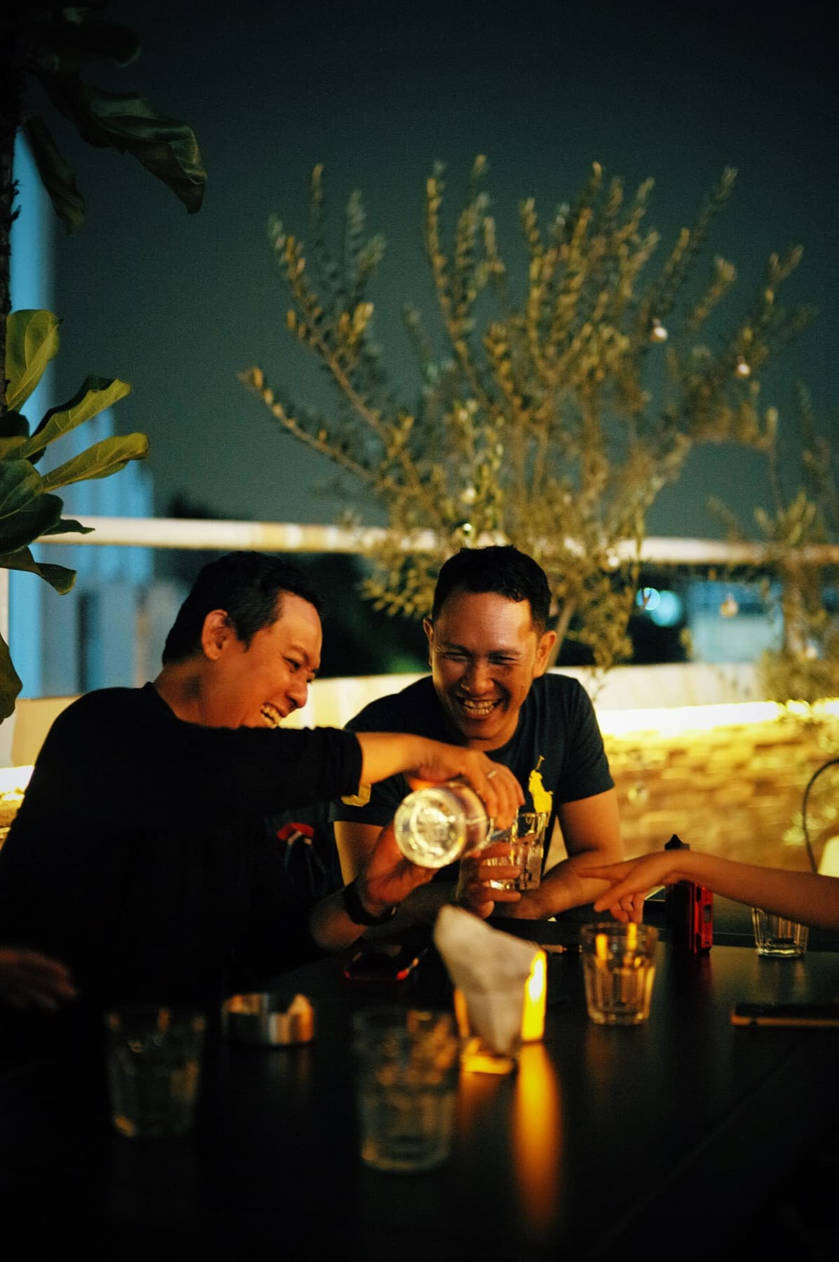 Two friends laughing and pouring a drink at an outdoor table at night